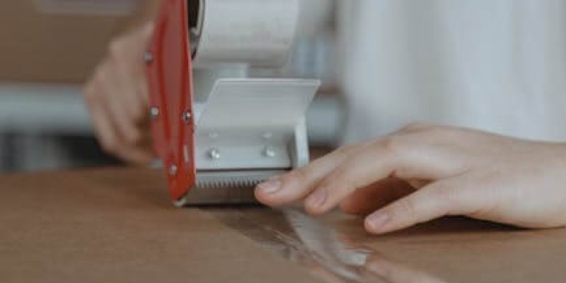 A person sealing a cardboard box with a tape dispenser in an indoor setting.